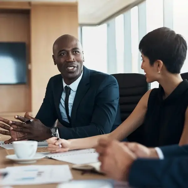 A man in a suit speaks and gestures while sitting at a conference table with colleagues, appearing engaged in a discussion about employee compensation. Notepads, documents, and coffee cups are spread out in the modern office setting.