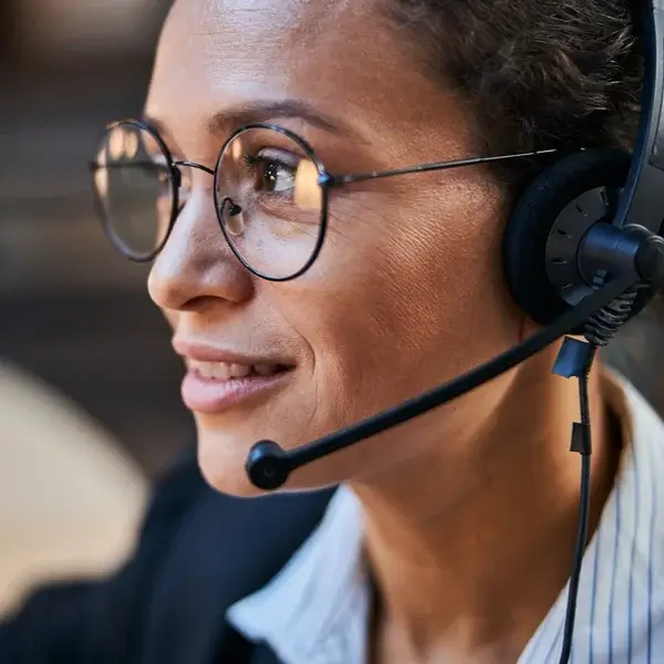 A woman wearing glasses and a headset with a microphone smiles while looking to the side, suggesting she is engaged in a conversation about employee benefits or providing customer service.