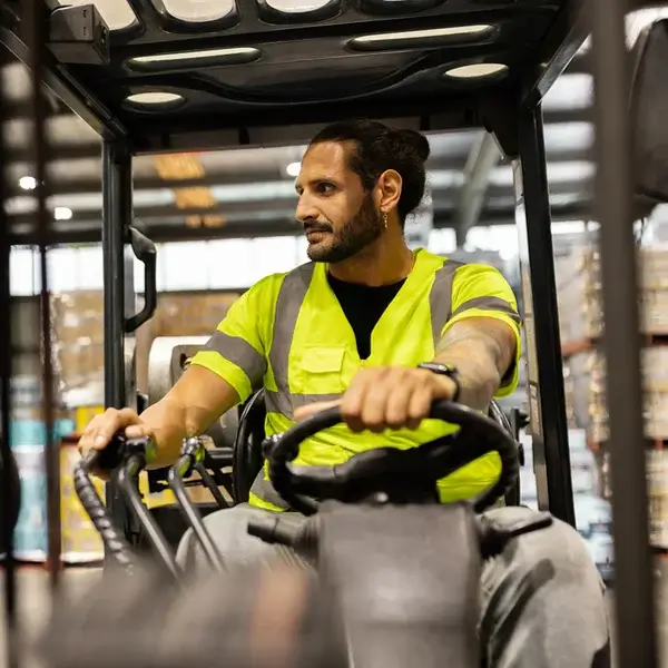 A man wearing a yellow safety vest operates a forklift inside a warehouse, looking to his right while holding the steering controls. Boxes and shelves are visible in the background, reflecting an organized approach to employee compensation management.