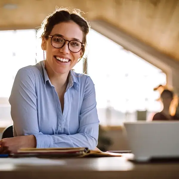 A woman wearing glasses and a light blue shirt smiles while sitting at a desk with papers and a cup in front of her, in a brightly lit room with large windows—discussing employee benefits and compensation.
