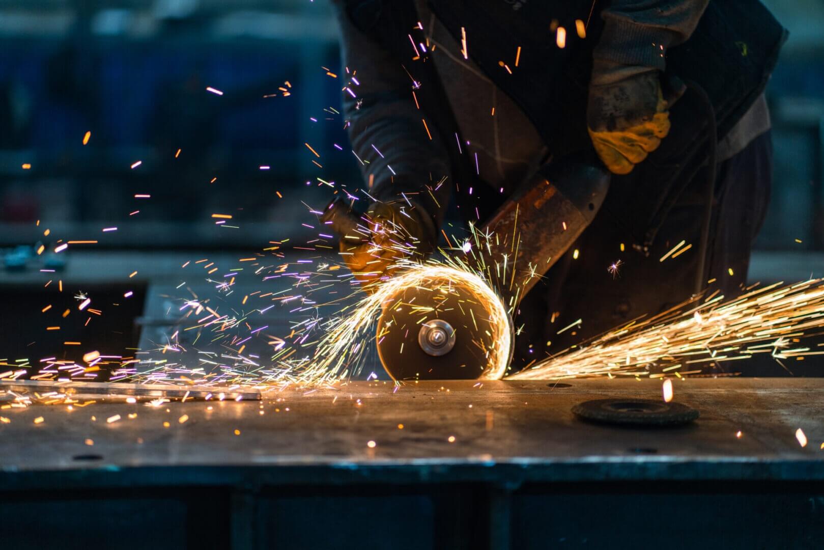 A worker using an angle grinder on a metal surface creates bright yellow sparks. Wearing gloves and protective clothing, they focus in a dimly lit workspace—a scene highlighting safety as vital as employee benefits in the workplace.