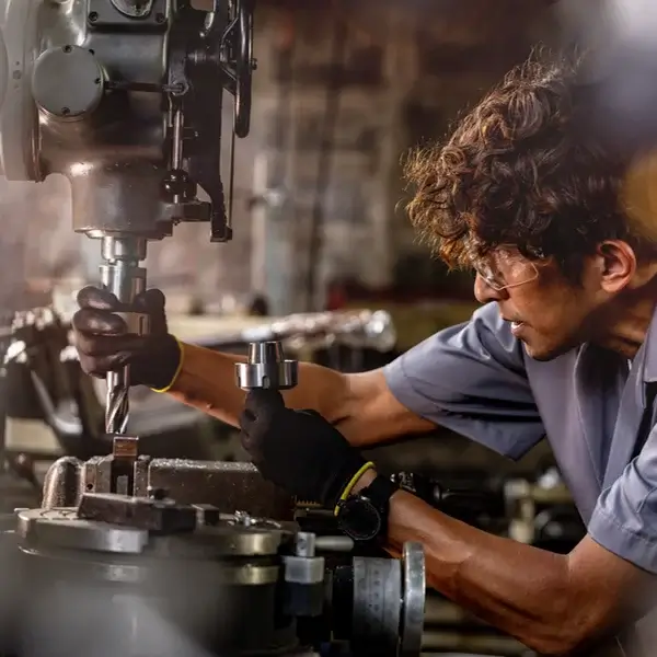 A person wearing safety glasses and gloves operates a large industrial drilling machine in a workshop, focusing closely on adjusting the equipment—part of a team committed to safety and fairness through strong compensation management.