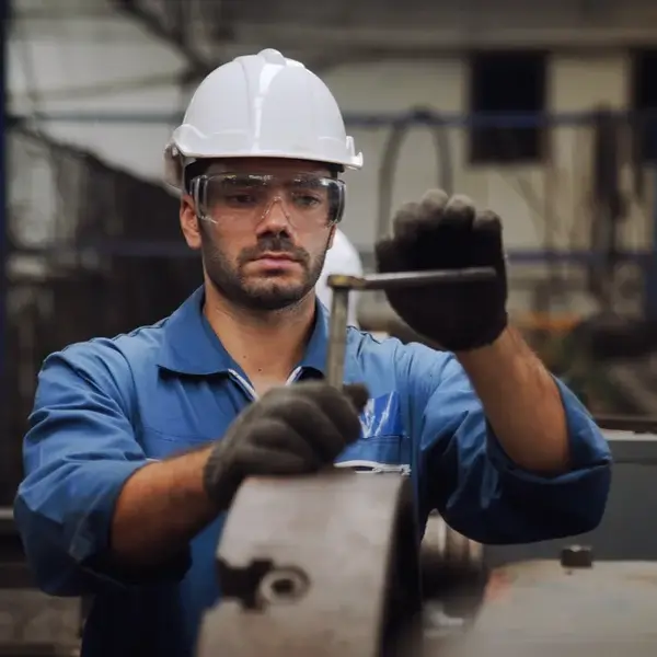 A man wearing a white hard hat, safety glasses, blue coveralls, and gloves is using a wrench to work on a large metal machine outdoors, highlighting the importance of employee compensation in demanding industrial roles.