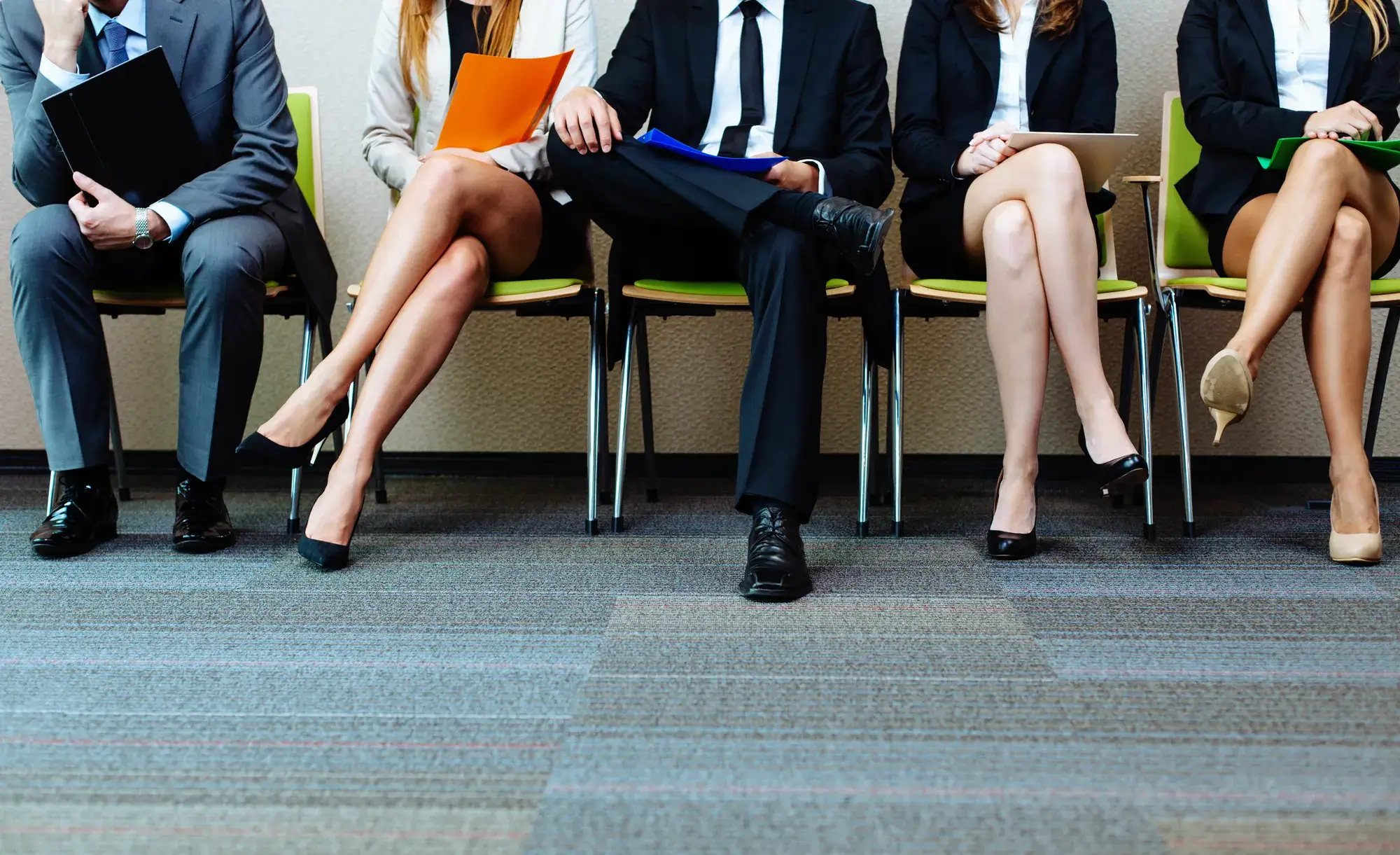 Four professionally dressed people sit in a row of chairs, each holding folders, their lower bodies and legs visible, suggesting they are waiting for an interview or meeting focused on salary benchmarking.