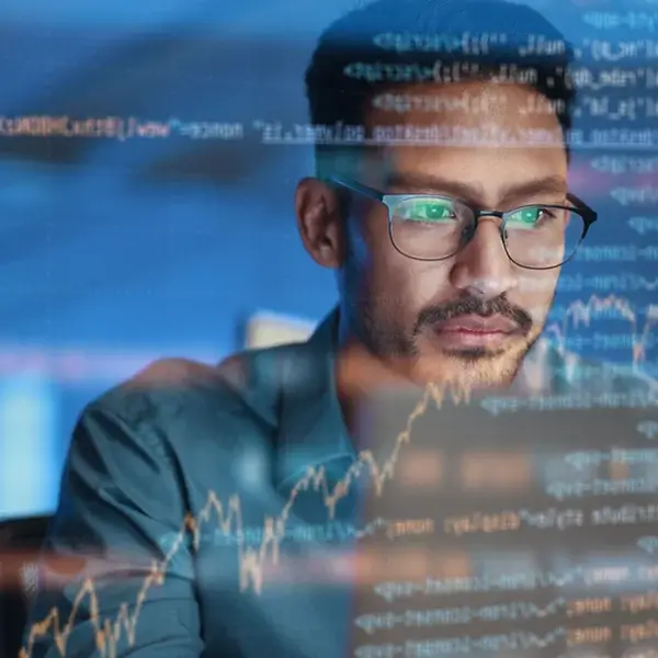 A man wearing glasses looks intently at a computer screen displaying lines of code and a rising stock chart, reflecting focus on compensation management and data analysis in a tech environment.